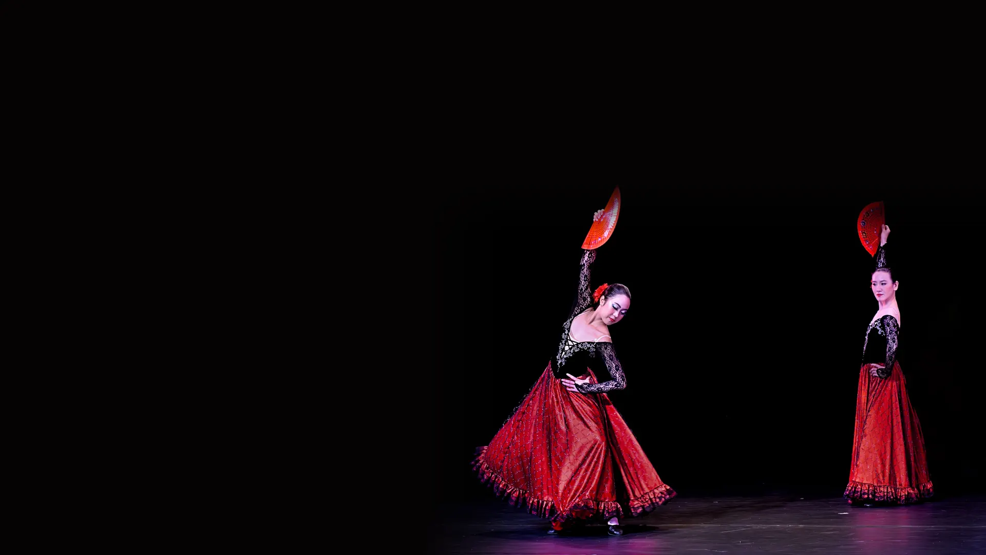 Two flamenco dancers in red ruffled skirts and black lace tops, each with a red fan, on a dark stage.