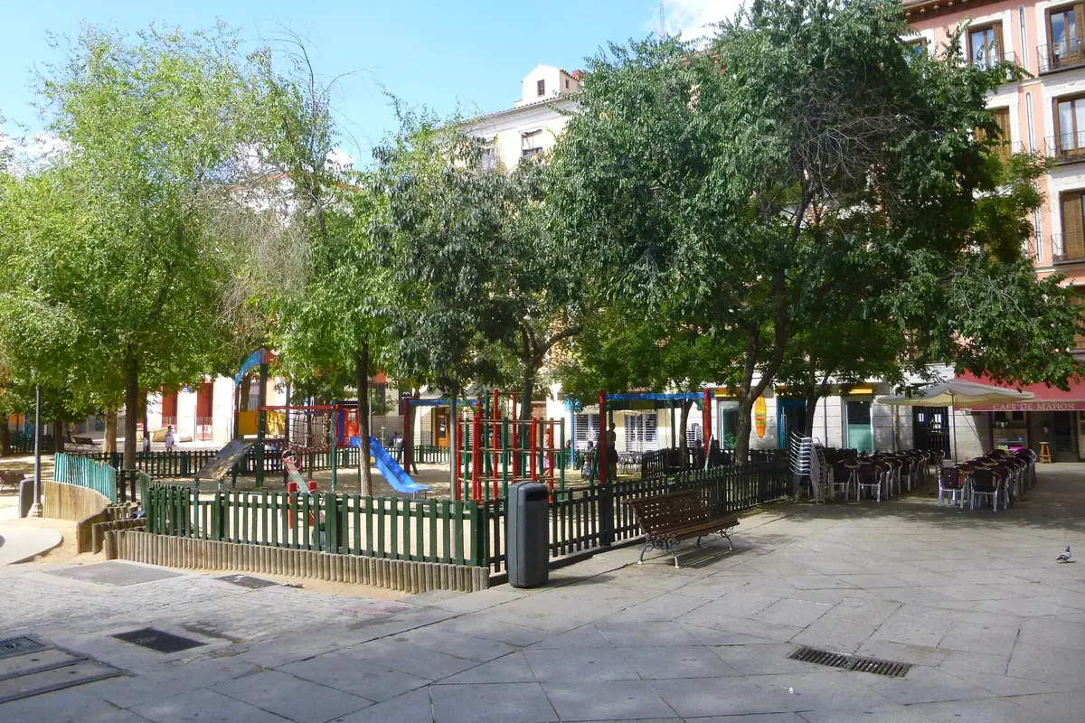A tree-shaded square with a children's playground, a bench and café tables under a blue sky.