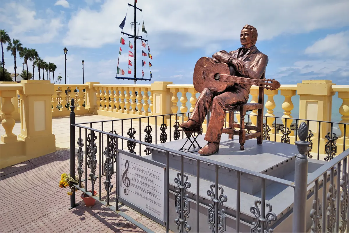 A bronze statue of a seated flamenco guitarist on a yellow balustraded terrace above the sea.